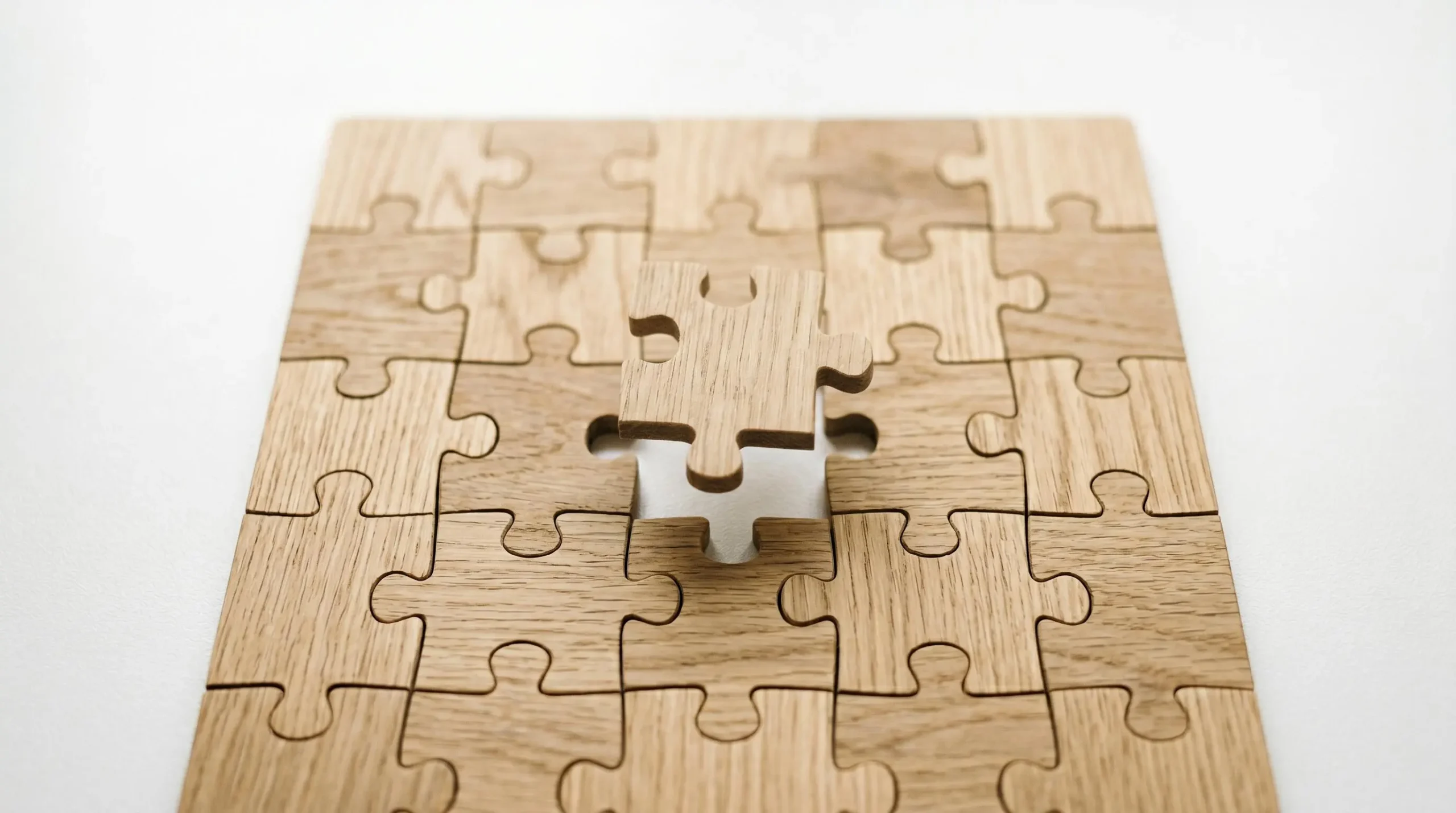 Pièces de puzzle en bois clair s'assemblant harmonieusement sur une surface blanche, vue de dessus, dernière pièce suspendue prête à compléter l'ensemble